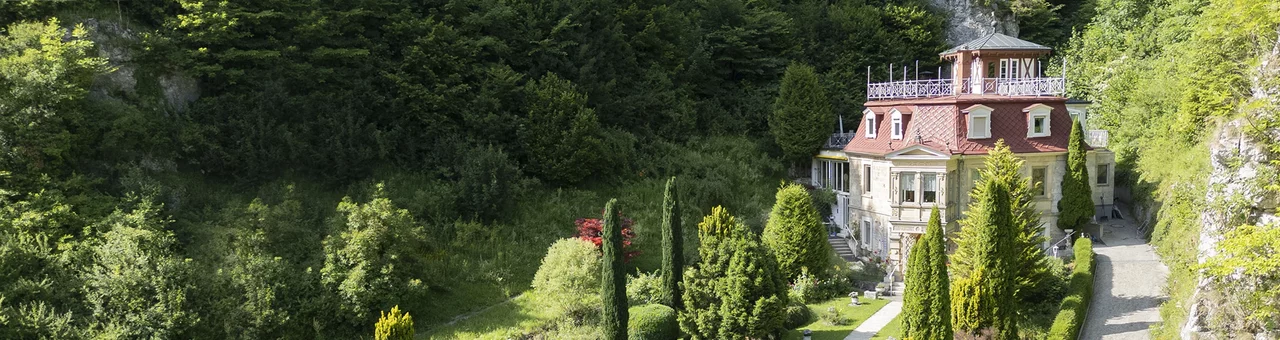 Panoramaaufnahme des SchlossFun, umgeben von bewaldeten Felsen, gepflegtem Garten und altem Baumbestand.